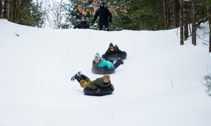 Three people wearing winter clothing are sliding down a snowy hill on inflatable tubes. A person stands at the top of the hill among trees.