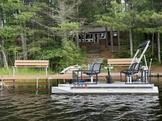 4013befa-84a7-45c8-a896-713f3eb10ee3 A small pontoon boat with two seats floats near a wooden dock and benches, with a cabin and steps surrounded by trees in the background.