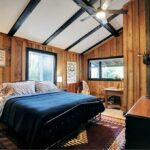 A cozy bedroom with wood-paneled walls, a large bed with a blue bedspread, two bedside lamps, a desk by the window, and a ceiling fan. Decorations include framed art and a deer head mount.