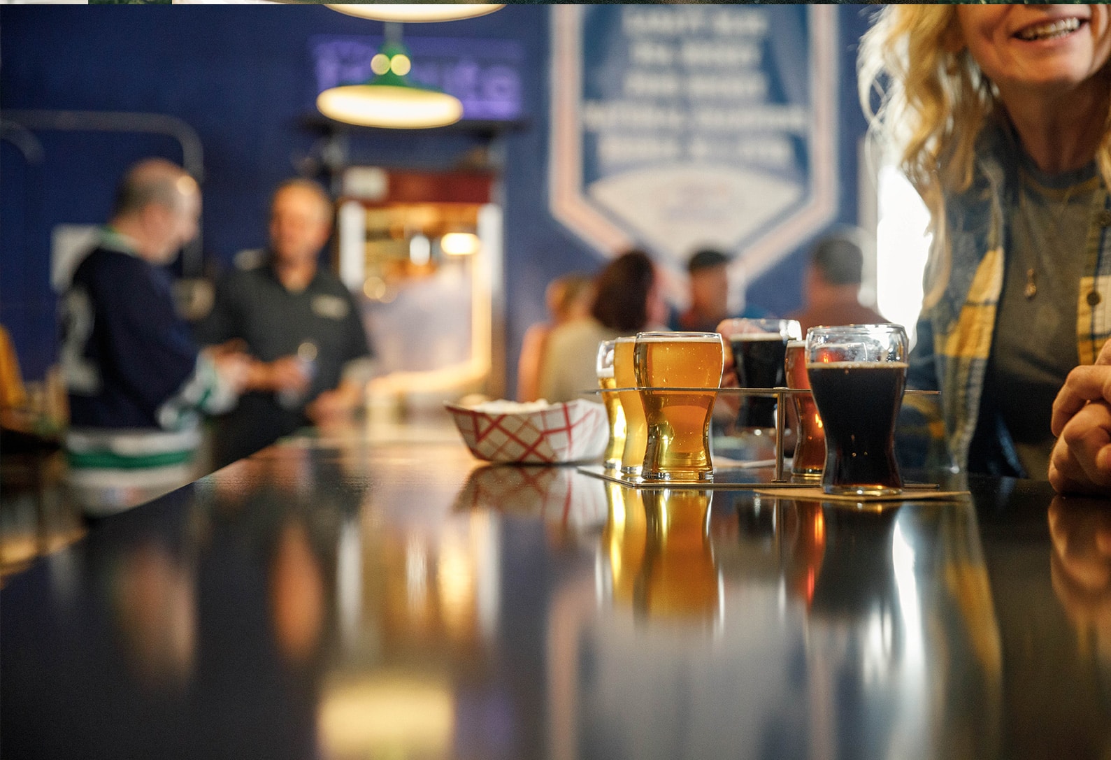 A variety of beers in glasses on a bar counter, with people in the background.