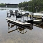 Small aluminum pontoon boat with two swivel chairs docked on a calm lake, surrounded by trees and a wooden bench on the dock nearby.