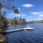 A white dock with a bench extends over a calm lake surrounded by trees under a partly cloudy blue sky.