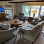 Living room with four gray armchairs around a circular wooden coffee table, floor lamp, side table, TV on wall, and large windows letting in natural light.