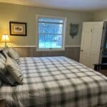 Bedroom with a large bed, plaid bedding, three pillows, a lamp on the nightstand, artwork on the wall, and a window letting in natural light. Door opens to an adjacent room.