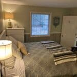 Bedroom with a bed covered in a sage green quilt and striped blankets. A lamp is on the bedside table, and a dresser with decor is against the wall. A window and partly open door are visible.