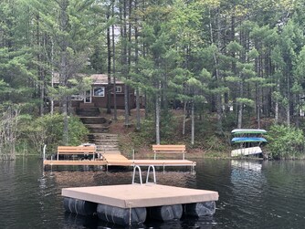 A wooden cabin is nestled among trees by a lake, with a dock extending into the water and a floating platform nearby. Canoes and kayaks are stacked on the shore.