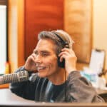 Person smiling while wearing headphones and speaking into a microphone in a recording studio.