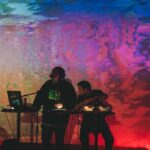 Two people operate electronic music equipment on stage with colorful, abstract lighting projected on the wall behind them.