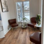 Small room with three brown leather chairs, two small white tables, a painting, and a water dispenser. A window shows snowy trees outside. The wood floor and white walls create a modern look.