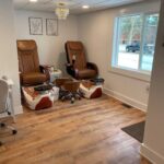 Two brown pedicure chairs with white foot basins in a salon room with wood flooring, white walls, framed pictures, a small chandelier, and a window near an open door.