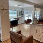 Interior of a hair salon with brown chairs, styling stations, mirrors, and hair products. A "welcome" sign is on the wall. Large windows provide natural light.