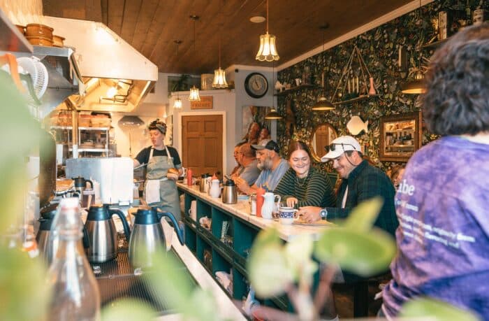 A group of people sit at a counter inside a cozy diner. A staff member in an apron works behind the counter. The ambiance is warm with decorative wallpaper and overhead lighting.