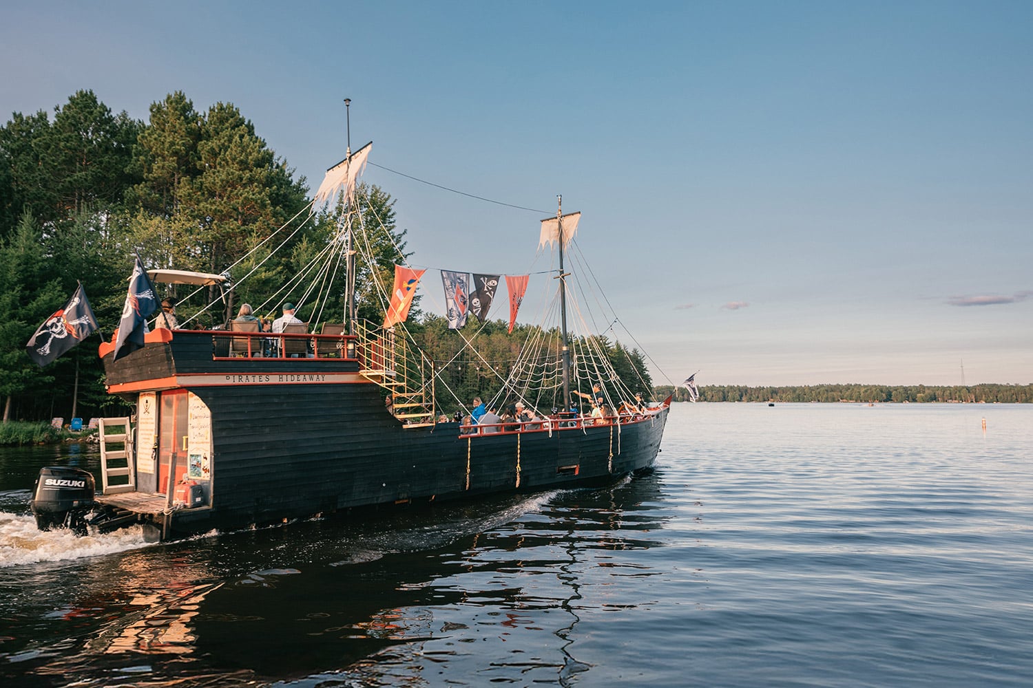 A large, black wooden ship with sails and pirate flags moves on a calm lake near a forested shoreline under a clear sky.