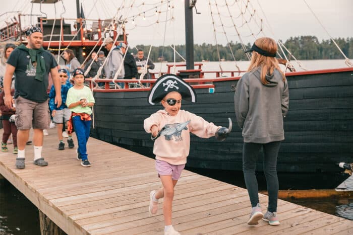 Children dressed as pirates walk and play on a wooden dock next to a large docked pirate ship, with adults and others in the background.