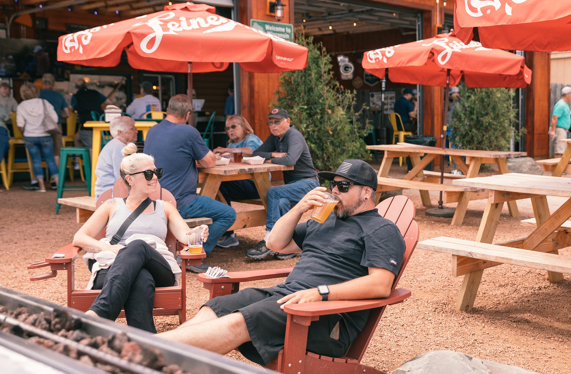 People sitting outdoors at a brewery, with red umbrellas overhead. A man drinks from a glass, while a woman holds a drink. Picnic tables are in the background.