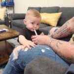 A baby sits on an adult’s lap, drinking from a straw held by the adult. Both are on a gray couch in a living room setting.