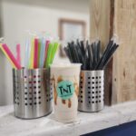 A plastic cup with a "TN Nutrition" logo, filled with a creamy drink and caramel drizzle, sits on a counter between two containers of colorful and black straws.