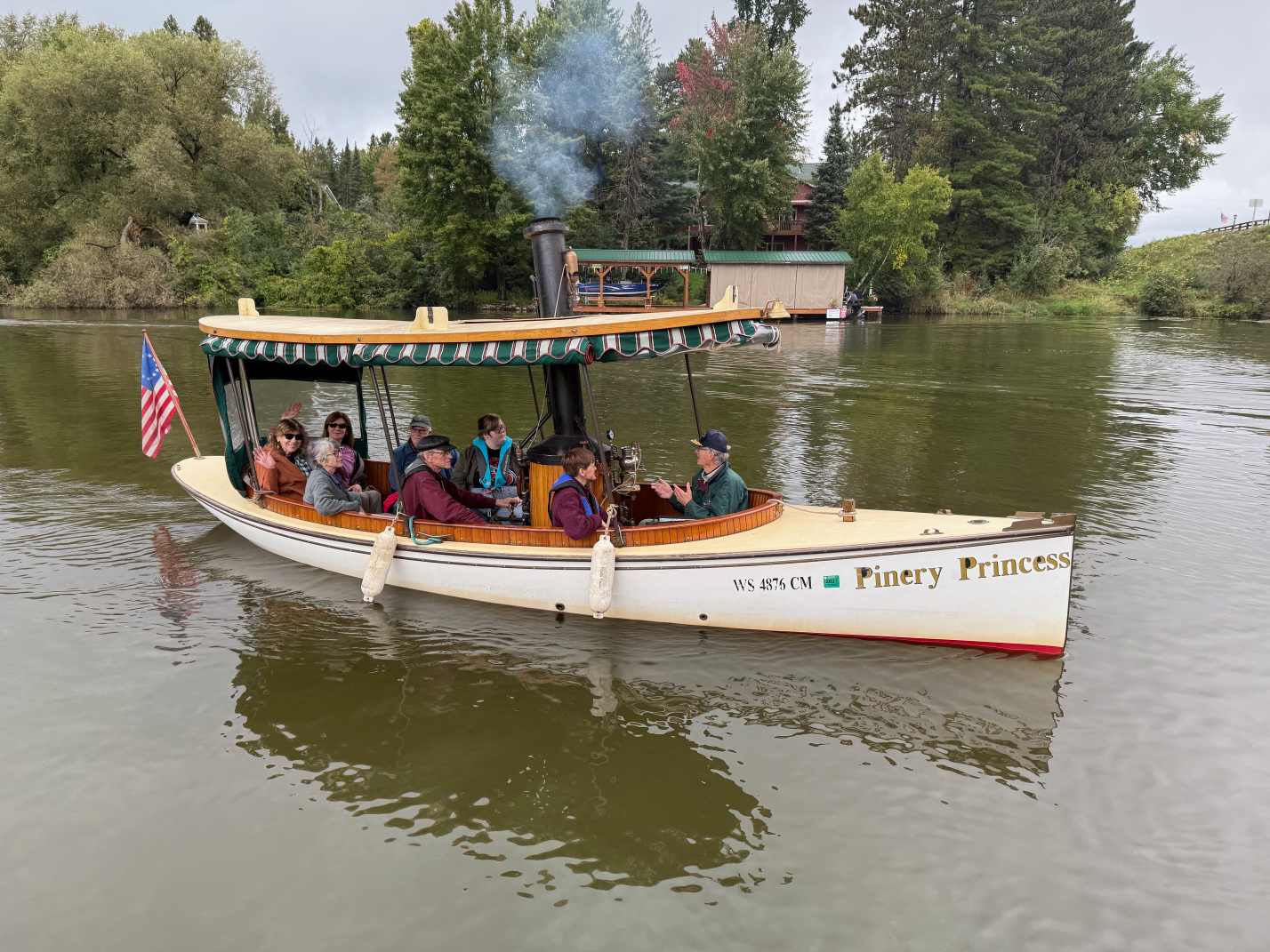 A small group of people rides on a white steam-powered boat named "Finery Princess" on a calm river, surrounded by trees and a dock in the background.