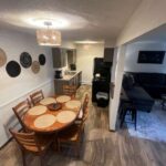 A dining area with a round wooden table set for six is adjacent to a kitchen and a living room with a dark sectional sofa and coffee table.
