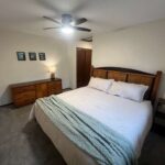 A bedroom with a large bed, wooden headboard, nightstand, dresser with a lamp, three wall pictures, and a ceiling fan.