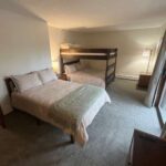 A bedroom with a double bed, a bunk bed, a floor lamp, a nightstand, and a sliding glass door letting in natural light.