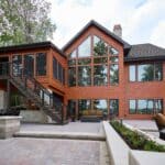 Modern two-story house with large glass windows, wooden siding, stone accents, upper-level deck, and spacious patio with outdoor seating surrounded by trees.