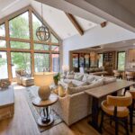 Open-concept living room and kitchen with large windows, vaulted ceiling, modern furnishings, a stone fireplace, and wooden accents throughout.
