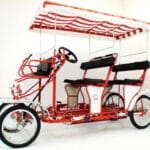 A four-wheeled, red and white pedal-powered surrey bike with a striped canopy and two bench seats, designed for multiple riders.