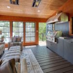 Modern sunroom with large windows, a grilling station, gray cabinetry, armchairs, a sofa, and a rug, overlooking a wooded lake view.