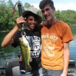 Two people on a boat, one holding up a largemouth bass with a fish gripper. They are smiling, with a lake and trees in the background.