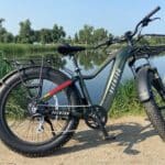 A black Aventon electric bicycle with thick tires is parked on a dirt path beside a calm lake, with greenery and trees in the background.