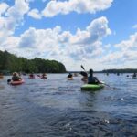 A group of people kayak on a calm lake under a partly cloudy sky, surrounded by trees along the shoreline.