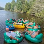 A group of people relax on green and yellow inflatable tubes in a river surrounded by trees on a sunny day.