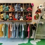 Wall cubbies hold rolled shirts in assorted colors above a row of hanging shirts; caps and hats are displayed on shelves and a step ladder nearby.