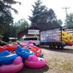 Brightly colored inflatable tubes are piled on the ground and loaded on a trailer beside a van, parked outside a rustic canoe outfitter building surrounded by trees.