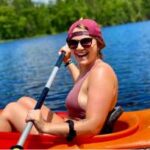 A person wearing sunglasses and a backwards cap paddles a red kayak on a lake surrounded by trees on a sunny day.