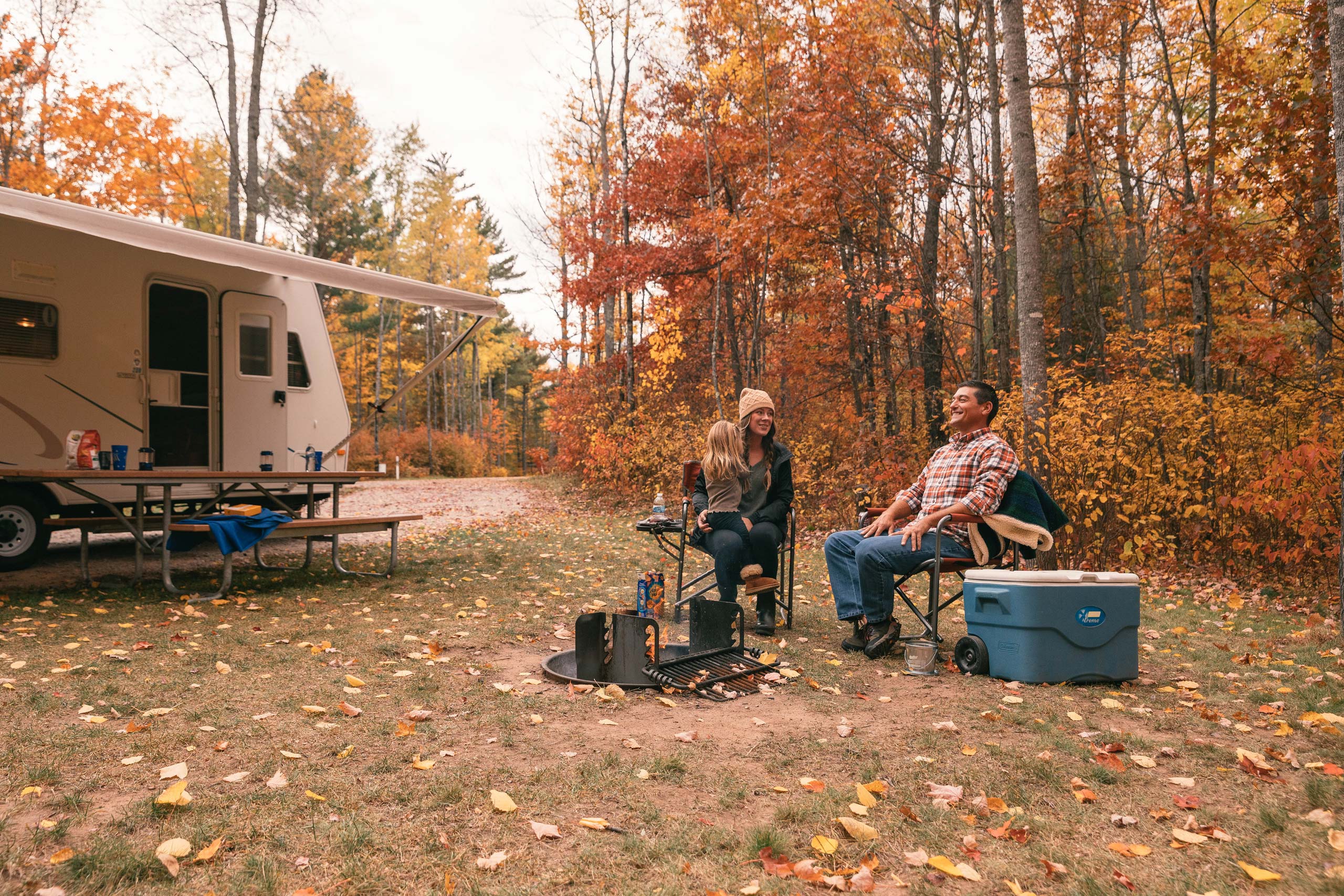 Two people sit by a campfire next to an RV and a blue cooler in a forest with autumn foliage. A picnic table and fire pit are also visible.
