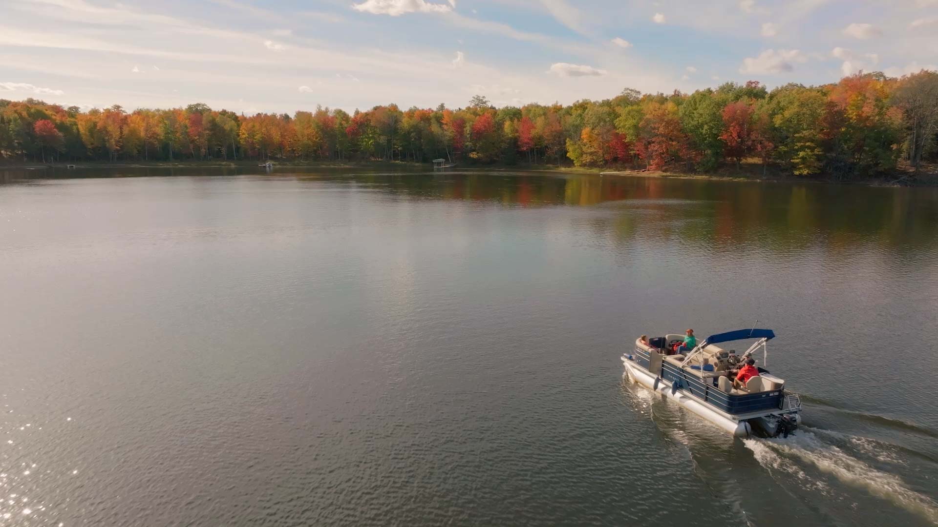 A pontoon boat with several people aboard moves across a calm lake, surrounded by trees with autumn foliage under a partly cloudy sky.