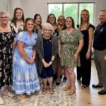 A group of nine women and one man stand together indoors, smiling at the camera. Some are wearing dresses, and one is in slacks and a polo shirt. Large windows are in the background.