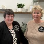 Two women wearing "True North Title" badges stand indoors in front of shelves with plants and decorative items, smiling at the camera.
