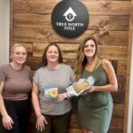 Three women stand in front of a "True North Title" sign; one holds a card with a sunflower, another holds a loaf of bread wrapped in foil.