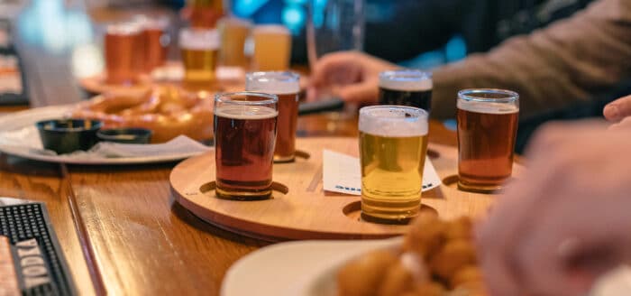 A wooden tray with four small glasses of beer sits on a bar table, surrounded by plates of food and people’s hands reaching for drinks.