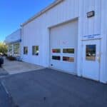 White metal building exterior with a garage door labeled "EXIT ONLY," several windows, and a door marked "Collision Center Office" on a paved surface under a clear blue sky.