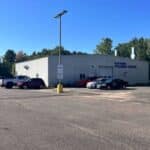 A metal building labeled "Parsons Collision Center" with several cars parked in front, surrounded by trees and under a clear blue sky.