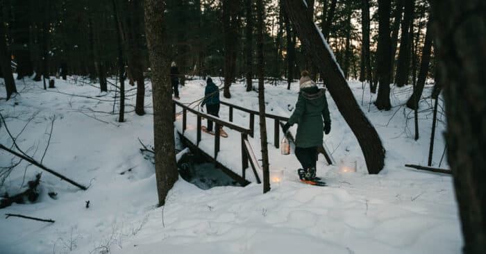 Two people in winter coats and snowshoes cross a small wooden bridge in a snowy forest, carrying lanterns in the late afternoon light.