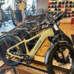 Rows of Aventon electric bikes displayed inside a bike shop, with helmets and accessories on the wall and bike gear visible in the background.