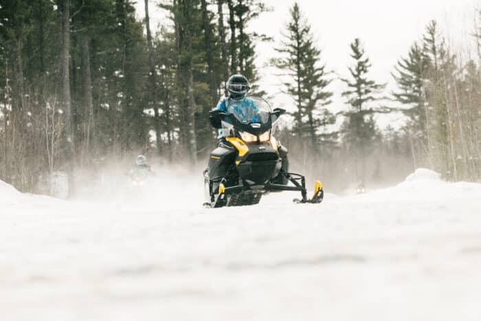 Person riding a yellow snowmobile on a snowy trail through a forest, with other snowmobiles visible in the background.