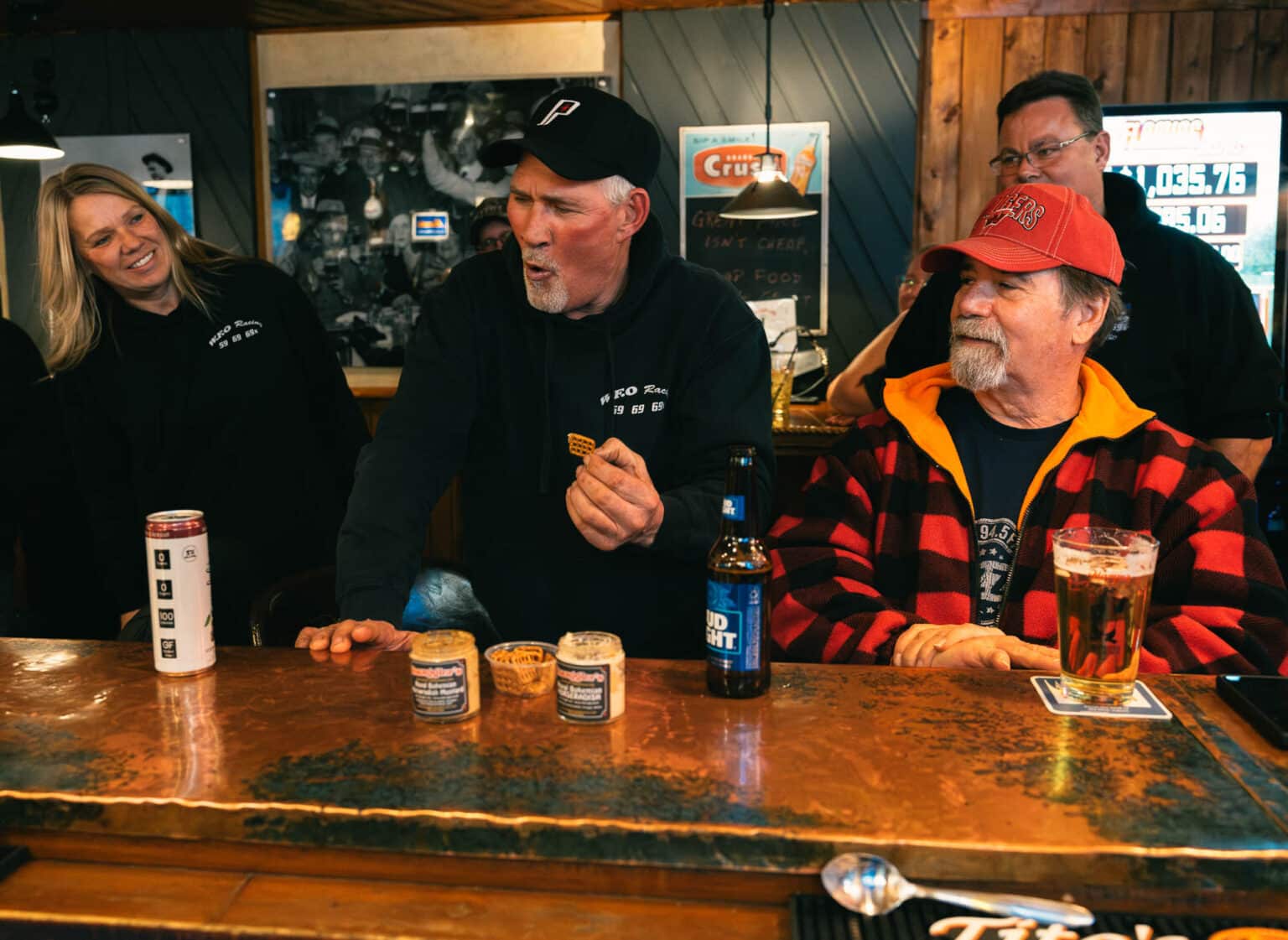 Four people sit at a bar; the man in the center, holding a snack, appears surprised while others look on. Drinks and jars of snacks are on the counter.