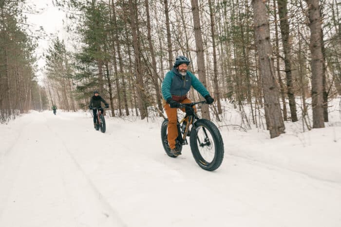Three people ride fat-tire bikes on a snowy trail through a forest of pine trees in winter.