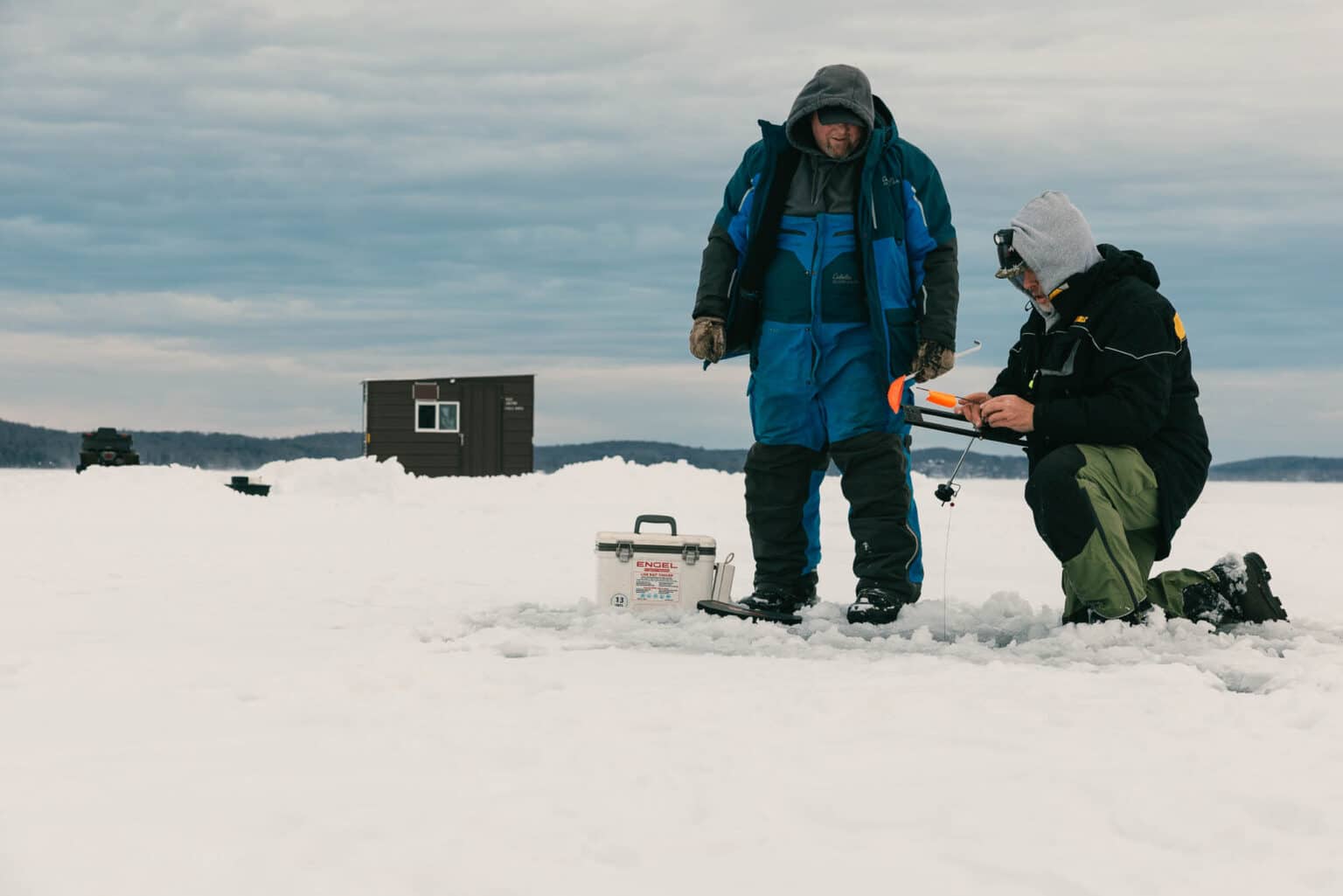 Two people dressed in winter clothing ice fishing on a frozen lake, with a small shelter and equipment visible in the background.